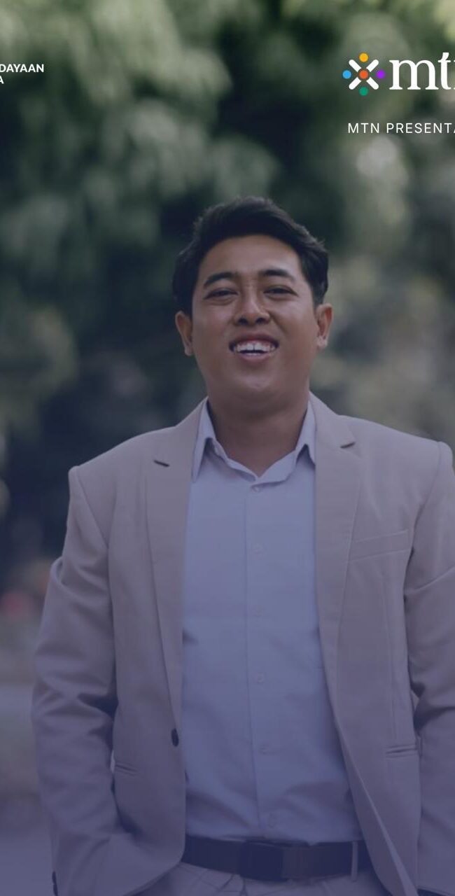 Alfa Zain mengenakan jas krem dan kemeja putih berpose dan tersenyum. Foto setengah badan.